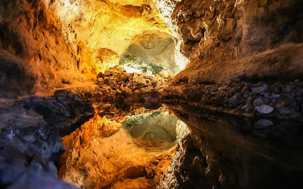 A lit lava tunnel with the rock roof reflected in still water on the floor