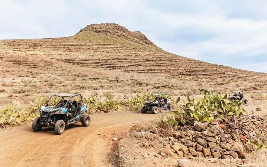 Two off-road buggies on a dirt track below a bare brown ridge under a wide sky