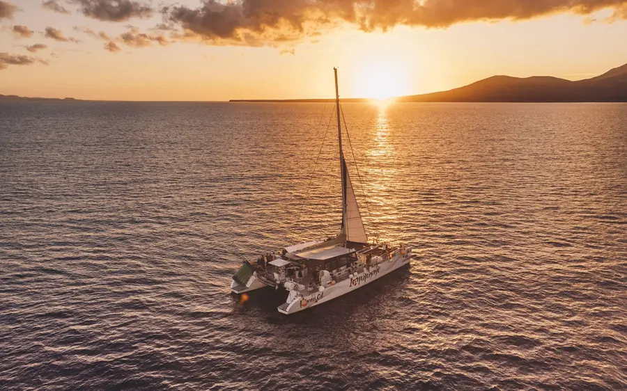 A catamaran on calm water with the sun setting behind a dark headland
