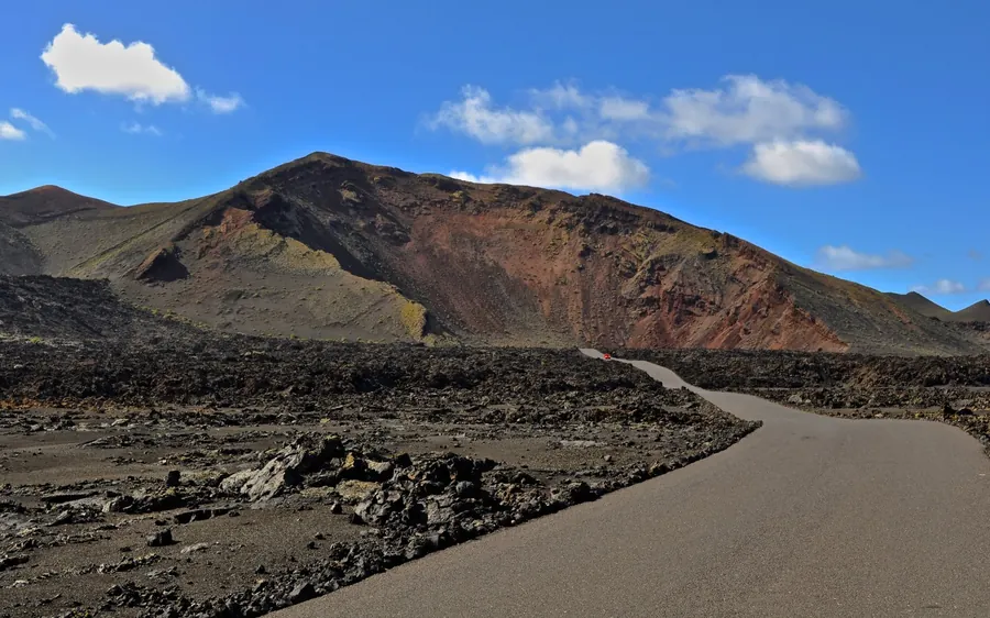 A smooth road curving between black lava and a dark cone under white cloud