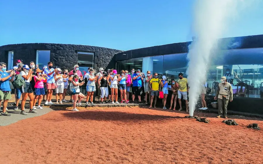 A crowd of visitors watching a tall plume of steam rise from the ground beside the park building