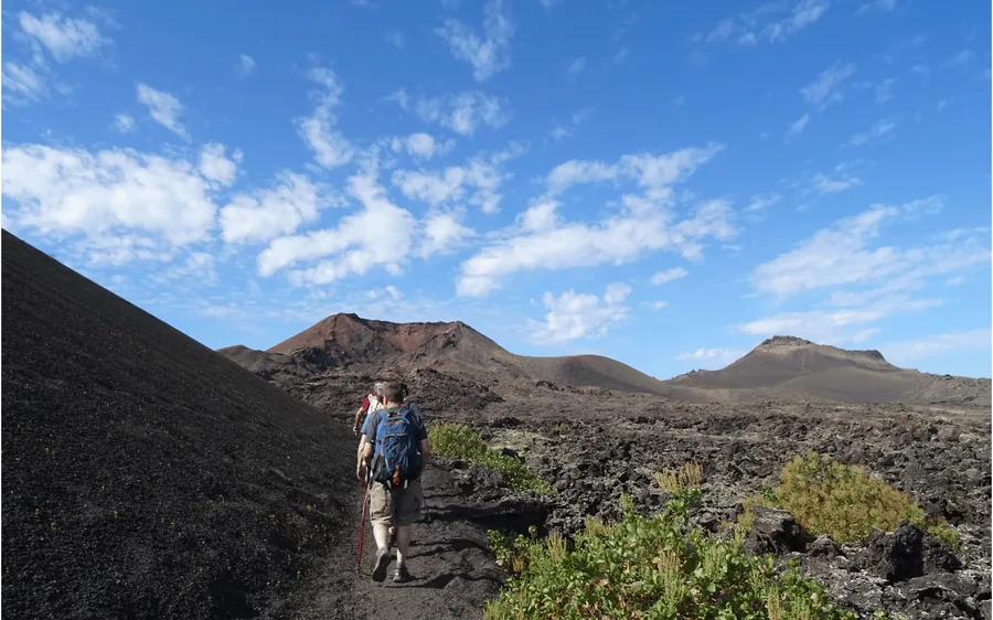 A walker with a rucksack on a path across a black cinder slope with cones ahead