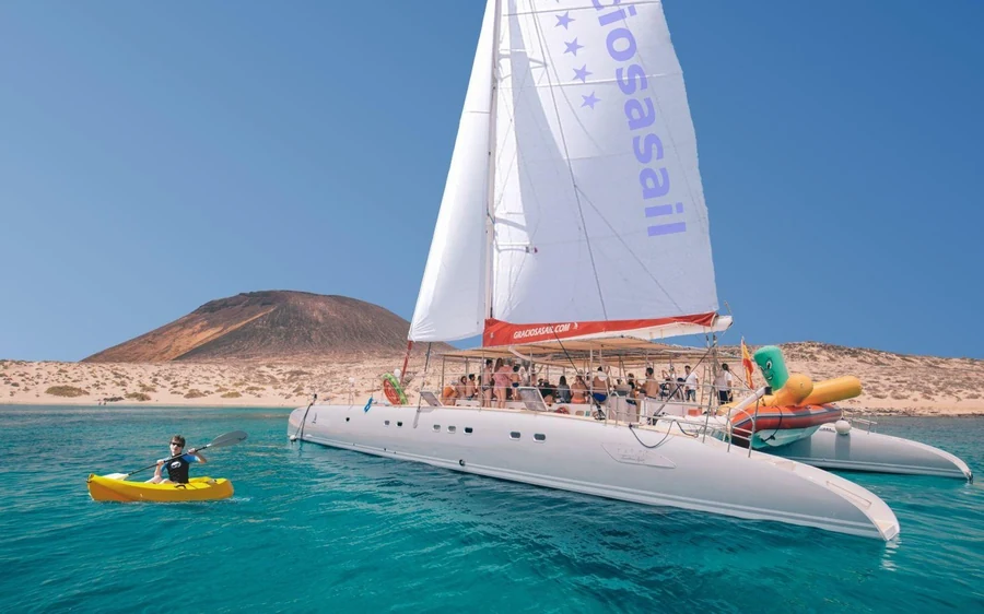 A white sailing catamaran off a pale beach with a volcanic cone behind and a kayaker alongside