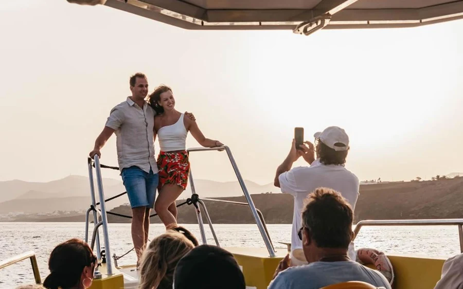 A couple standing at the rail of a boat at sunset while another passenger photographs them
