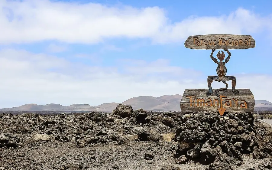 The Timanfaya park sign standing on broken black lava with low hills behind